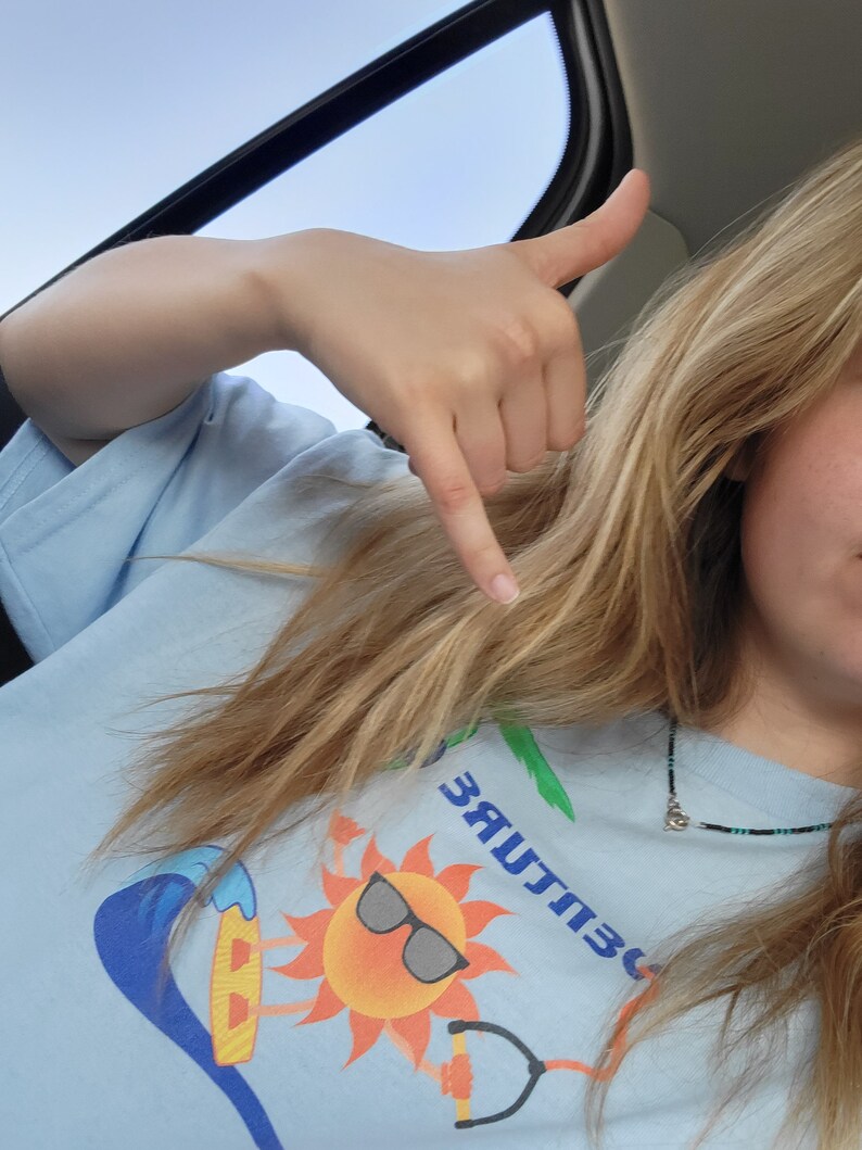 May include: A person wearing a light blue t-shirt with a graphic of a sun wearing sunglasses and riding a surfboard on a wave. The text "303 CULTURE" is printed on the shirt.