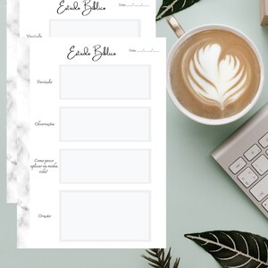 Pode incluir: Duas páginas brancas de estudo bíblico com o título "Estuda Biblica" e seções para versículos, observações e oração. Uma xícara de café com arte latte, um teclado e vegetação também estão na imagem.
