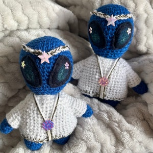 Op de afbeelding: Twee handgemaakte blauwe gehaakte alienpoppen met grote ogen en steraccenten. Ze dragen witte jasjes met decoratieve kettingen en liggen op een zachte, grijze deken. De poppen hebben een grillig, handgemaakt uiterlijk.