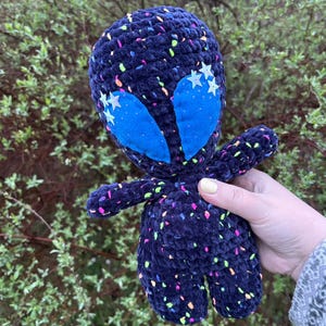 Puede incluir: Una muñeca alienígena de felpa hecha a mano, en hilo azul oscuro con motas coloridas. El alienígena tiene ojos azules grandes y detalles en forma de estrella. La muñeca está sostenida en la mano de una persona.