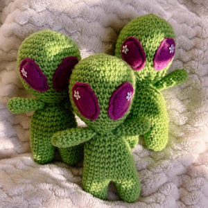 Op de afbeelding: Drie handgemaakte groene gehaakte alienpoppen met grote paarse ogen en kleine witte bloemaccenten. De poppen hebben kleine armen en benen en zijn geplaatst op een zacht, getextureerd wit oppervlak. De aliens zijn een grillig, handgemaakt item.
