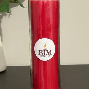 May include: A tall, cylindrical red wax candle encased in a clear glass jar. A circular white label is affixed to the centre of the glass, featuring a graphic of a lit candle flame above the text FJM CANDLES in a dark brown serif font.