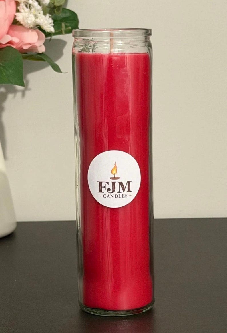 May include: A tall, cylindrical red wax candle encased in a clear glass jar. A circular white label is affixed to the centre of the glass, featuring a graphic of a lit candle flame above the text FJM CANDLES in a dark brown serif font.