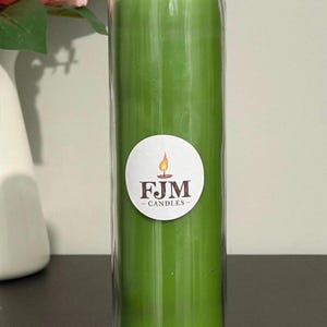 May include: A tall, cylindrical green candle encased in a clear glass jar. A circular white label is centred on the front of the glass, featuring a graphic of a lit flame above the dark brown text FJM CANDLES in a serif font.