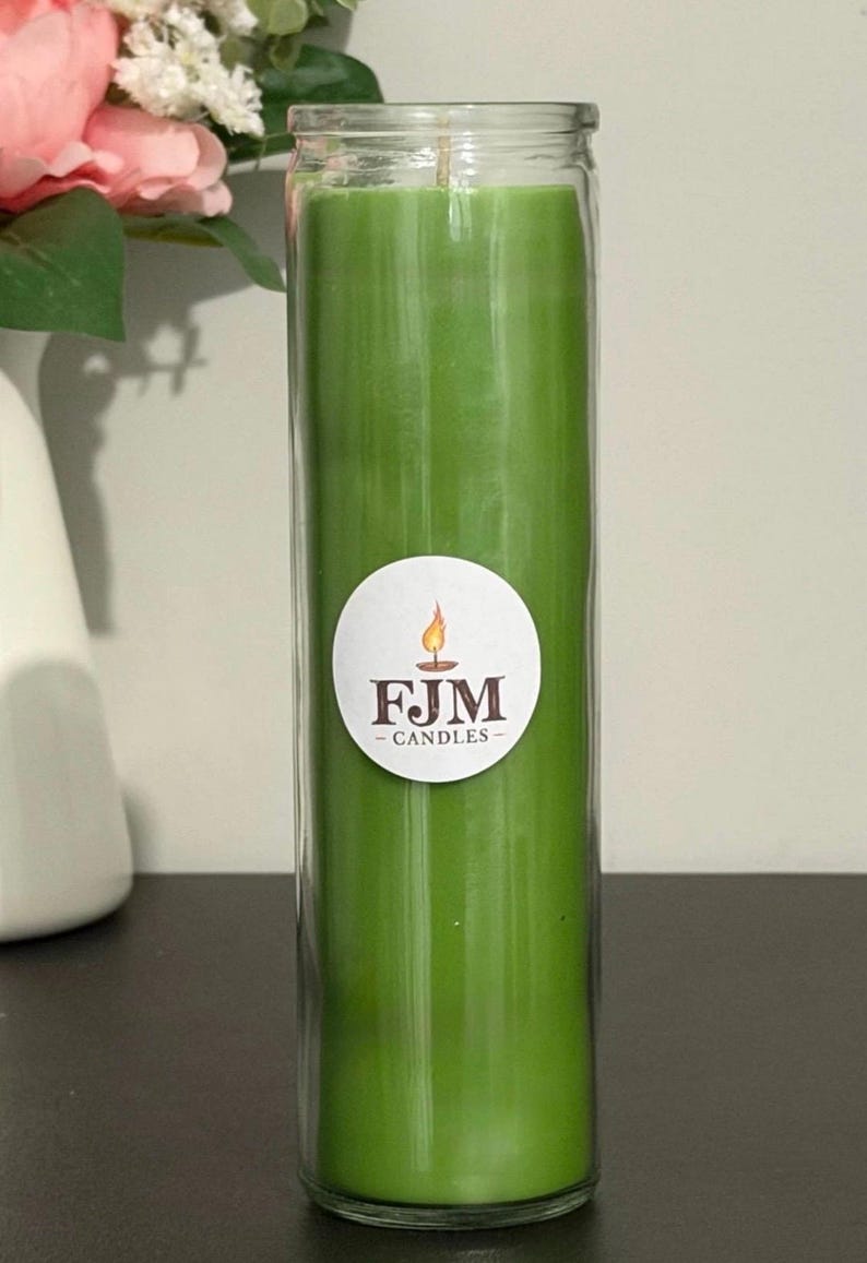 May include: A tall, cylindrical green candle encased in a clear glass jar. A circular white label is centred on the front of the glass, featuring a graphic of a lit flame above the dark brown text FJM CANDLES in a serif font.