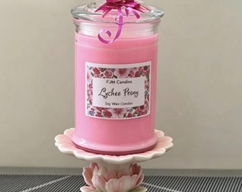 May include: A pink soy wax candle in a clear glass jar with a lid, topped with a magenta ribbon. The label reads FJM Candles Lychee Peony Soy Wax Candles. The jar sits on a ceramic pedestal stand shaped like a pink and green lotus flower.