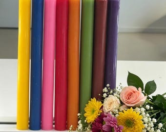 May include: A set of eight tall, smooth, cylindrical taper candles in vibrant colors including yellow, blue, pink, red, orange, green, brown, and purple, arranged in a row next to a bouquet of mixed roses and daisies with baby's breath.