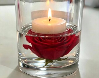 May include: A clear glass cylinder filled with water containing a single vibrant red rose blossom submerged beneath a white tea light candle. The candle is lit, casting a warm glow, and the rose petals are fully visible through the transparent glass.