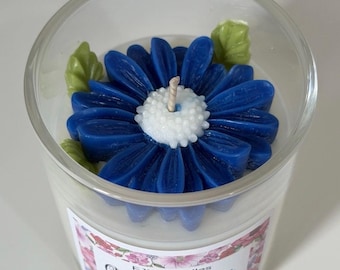 May include: A clear glass jar candle containing white soy wax topped with a blue flower-shaped wax decoration featuring a white center and green leaves. The label reads FJM Candles Ocean Breeze Soy Wax Candles with a floral border design.