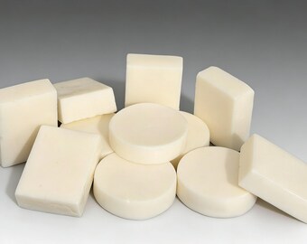 May include: A collection of ivory-colored soaps in various shapes, including rectangular bars, square blocks, and round discs. The soaps are arranged in a pile, with some overlapping. The lighting is soft, highlighting the smooth surfaces.