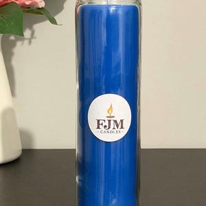 May include: A tall, cylindrical blue candle encased in a clear glass jar. A circular white label is centred on the front of the glass, featuring a graphic of a lit candle flame above the dark brown text FJM CANDLES in a serif font.
