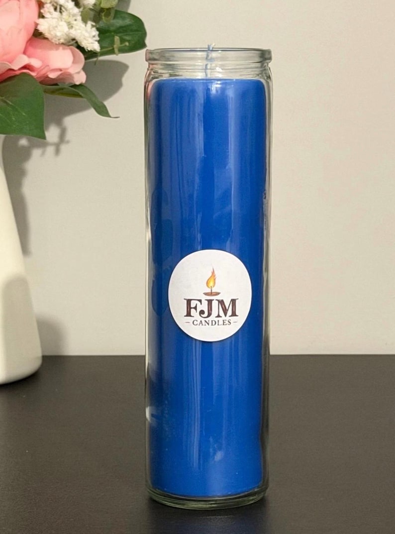 May include: A tall, cylindrical blue candle encased in a clear glass jar. A circular white label is centred on the front of the glass, featuring a graphic of a lit candle flame above the dark brown text FJM CANDLES in a serif font.