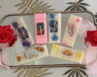 May include: An arrangement of wax melts in various colors and scents, including lilac, bubblegum, and baby powder, displayed on a mirrored tray. The melts are rectangular and labeled. Red roses and ribbons decorate the tray.