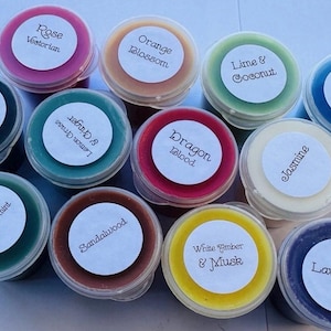 Puede incluir: Una colección de doce recipientes redondos de plástico con ceras aromáticas de colores. Cada tapa tiene una etiqueta blanca circular con texto negro que identifica aromas como Rose Victorian, Orange Blossom, Lime & Coconut, Rio De Janeiro, Black Raspberry Sugar, Lemongrass & Ginger, Dragon Blood, Jasmine, Eucalyptus & Spearmint, Sandalwood, White Amber & Musk y Lavender.