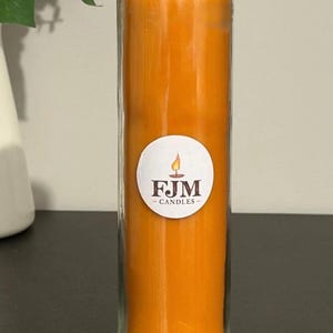 May include: A tall, cylindrical glass candle filled with solid orange wax. A circular white label is centred on the front of the glass, featuring a small flame graphic above the dark brown text FJM CANDLES in a serif font.
