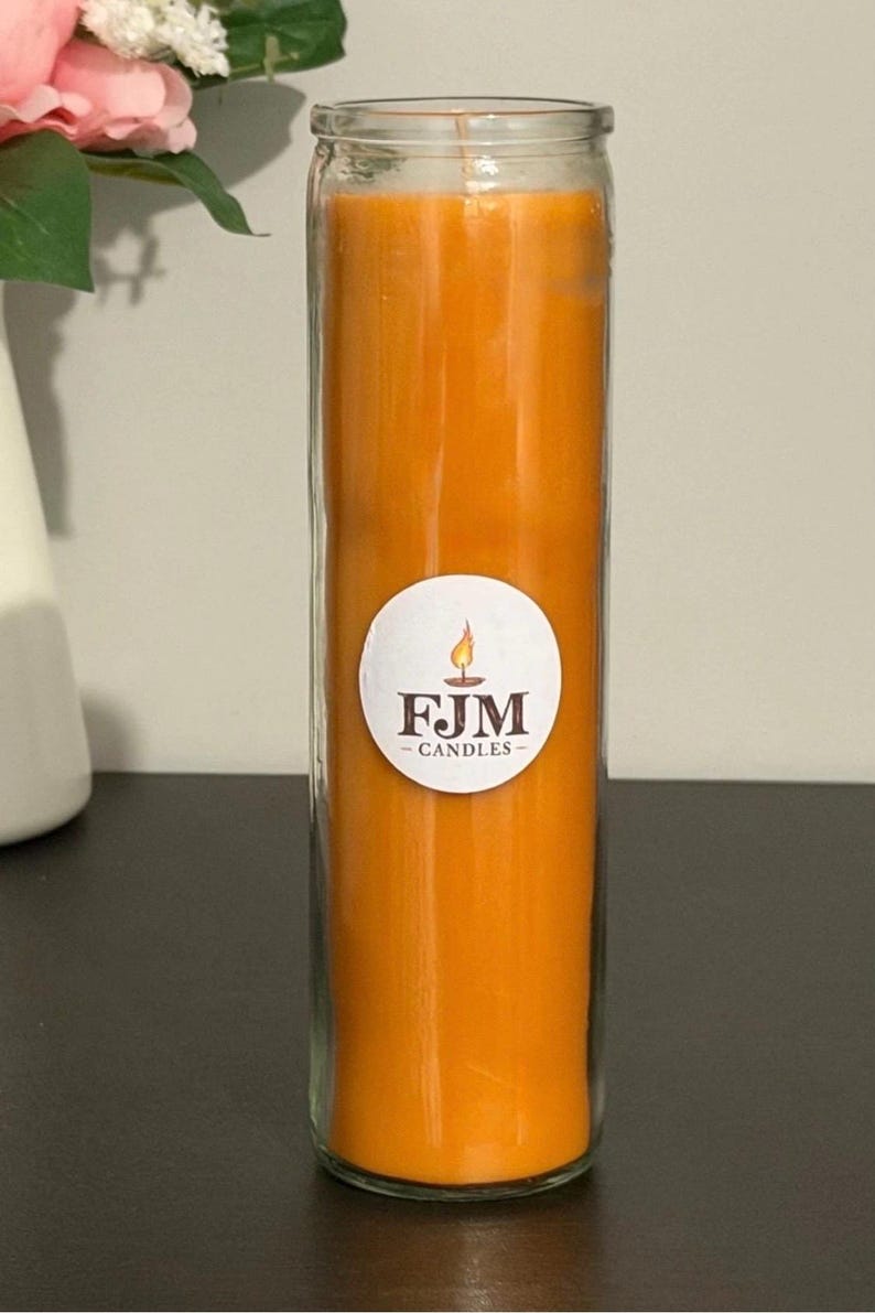 May include: A tall, cylindrical glass candle filled with solid orange wax. A circular white label is centred on the front of the glass, featuring a small flame graphic above the dark brown text FJM CANDLES in a serif font.