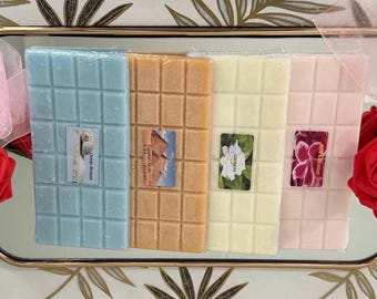 May include: Four rectangular wax melt bars in light blue, tan, cream, and pale pink, each divided into small squares. Each bar features a small rectangular label with imagery and text, arranged on a gold-rimmed tray with red roses.