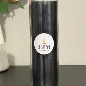 May include: A tall, cylindrical glass candle holder filled with solid black wax. A circular white sticker is centred on the glass, featuring a graphic of a lit candle and the text FJM CANDLES in dark brown lettering with a small flame icon above.