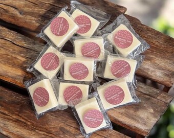 May include: A collection of ten rectangular bars of off-white soap, each individually wrapped in clear plastic. Every bar features a circular maroon sticker printed with the text Handmade Soap Travel Size. The bars are arranged in a cluster.