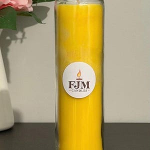 May include: A tall, cylindrical glass candle filled with bright yellow wax. A circular white label is centred on the glass, featuring a small flame icon above the dark brown text FJM CANDLES. The candle has a single visible wick at the top.