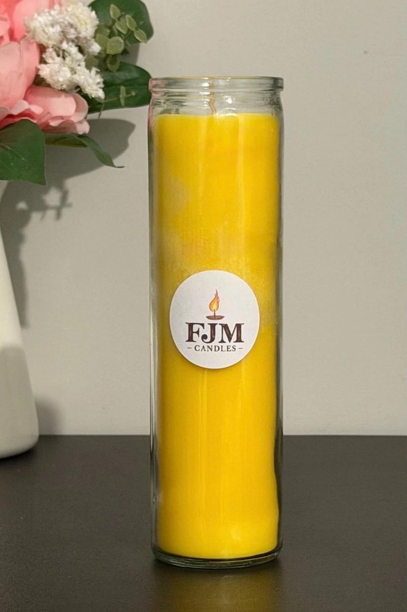 May include: A tall, cylindrical glass candle filled with bright yellow wax. A circular white label is centred on the glass, featuring a small flame icon above the dark brown text FJM CANDLES. The candle has a single visible wick at the top.