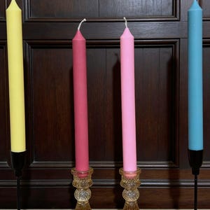 May include: Four tall taper candles in yellow, dark pink, light pink, and light blue. The yellow and light blue candles are held in thin black metal stands, while the dark pink and light pink candles are in amber-coloured glass candle holders.