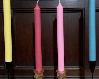 May include: Four tall taper candles in yellow, dark pink, light pink, and light blue. The yellow and light blue candles are held in thin black metal stands, while the dark pink and light pink candles are in amber-colored glass candle holders.