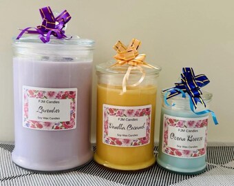 May include: Three glass jar soy wax candles of varying sizes. The largest is light purple labeled Lavender, the middle is yellow labeled Vanilla Caramel, and the smallest is light blue labeled Ocean Breeze. Each has a floral label and a decorative bow.
