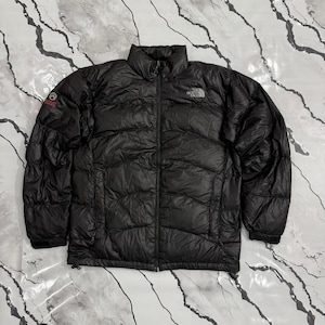 Könnte beinhalten: Schwarze Steppjacke mit durchgehendem Reißverschluss und hohem Kragen. Das The North Face-Logo ist auf der Brust zu sehen, und ein Summit Series-Logo befindet sich auf dem linken Ärmel. Die Jacke ist aus leichtem Material.