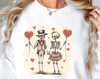 May include: White sweatshirt featuring a whimsical illustration of two skeleton figures holding hands and heart-shaped balloons. The male skeleton wears a top hat and suit, while the female skeleton wears a skirt. Red hearts are scattered around the figures.