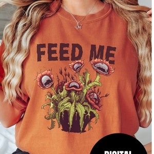 May include: A rust-colored t-shirt with the text "FEED ME" printed in black. The shirt features a graphic of a carnivorous plant with multiple mouths.