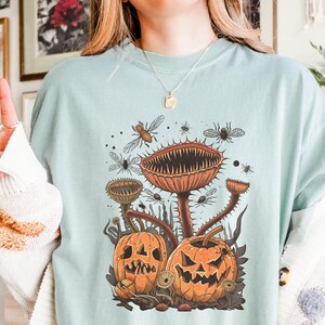 Op de afbeelding: Lichtblauw T-shirt met een Halloween-thema. Het ontwerp bevat twee uitgesneden pompoenen, een grote paddenstoel en verschillende insecten. De tekst "ALL HANDY PRINTABLE" staat rechtsonder.