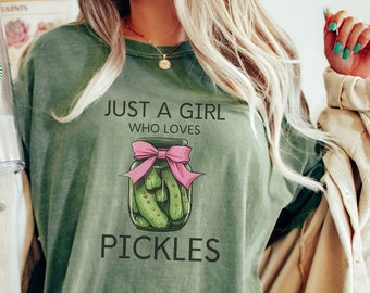 May include: Olive green t-shirt with the text "JUST A GIRL WHO LOVES PICKLES." The graphic features a jar of pickles tied with a pink bow. The shirt is a relaxed fit.