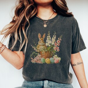 Puede incluir: Una camiseta gris oscuro con una ilustración vintage de un conejo marrón sentado en una maceta de flores con huevos de Pascua coloridos. La camiseta tiene cuello redondo y mangas cortas.