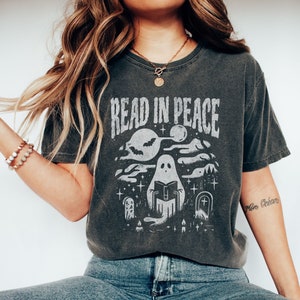 May include: A dark gray t-shirt with a white graphic of a ghost reading a book in a graveyard setting. The text "Read in Peace" is above the ghost.