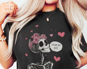 May include: Black t-shirt featuring a skeleton graphic wearing a floral crown and surrounded by pink hearts. The skeleton has a speech bubble that says "You're my Boo". A fun and spooky design for Halloween or everyday wear.