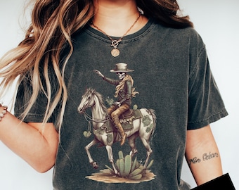 May include: A dark gray t-shirt with a graphic of a skeleton riding a horse through a desert landscape. The skeleton is wearing a hat and holding a gun. The horse is white with brown spots and has a saddle. The background is a desert landscape with cacti.