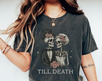 May include: A dark gray t-shirt with a graphic of two skeletons dressed as a bride and groom. The skeletons are holding a bouquet of roses and the text "Till Death" is printed below them.