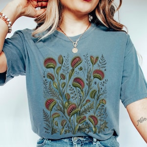Puede incluir: Una camiseta azul de manga corta con una ilustración botánica detallada de varias plantas atrapamoscas con hojas verdes y rojo púrpura, intercaladas con varios follajes tipo helecho y pequeñas arañas sobre un fondo claro.