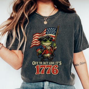 Könnte beinhalten: Ein anthrazitfarbenes T-Shirt mit einer Grafik eines Frosches in militärischer Kleidung des 18. Jahrhunderts, der eine Sonnenbrille trägt und eine amerikanische Flagge hält. Unter dem Frosch steht der Text: OFF TO ACT LIKE IT'S 1776 in fetten gelben und roten Buchstaben.