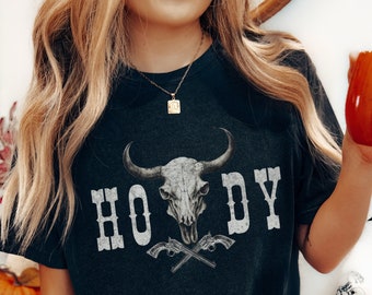 Könnte beinhalten: Dunkelgraues T-Shirt mit einem Stierschädel-Grafik und dem Wort "HOWDY" in weißen Blockbuchstaben. Unter dem Text befinden sich zwei gekreuzte Pistolen. Die Worte "DIGITALER DOWNLOAD" stehen in einem schwarzen Kreis unten.