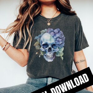 May include: A dark grey t-shirt featuring a blue and white skull graphic. The skull is surrounded by blue and green flowers. The text "Mère Océan" is visible on the right sleeve of the shirt.