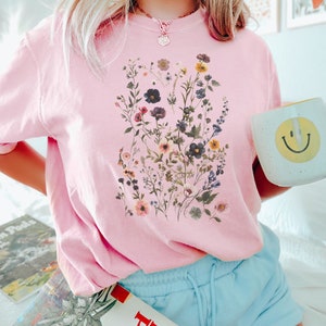 May include: A pink t-shirt with a floral print featuring a variety of flowers and greenery. The shirt has a crew neck and short sleeves.