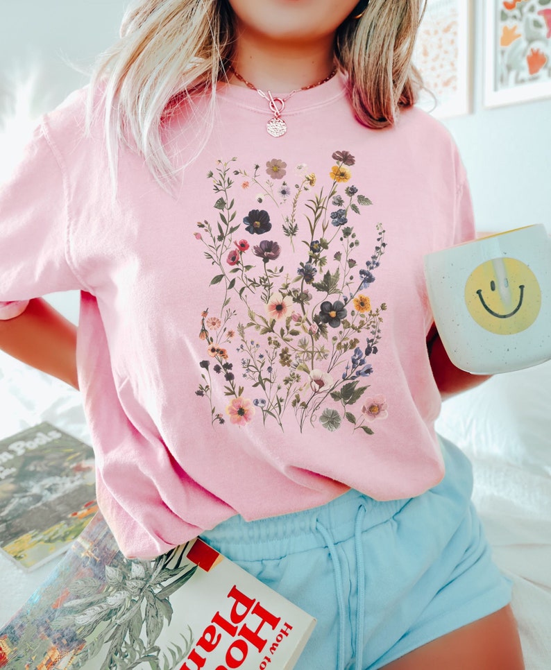 May include: A pink t-shirt with a floral print featuring a variety of flowers and greenery. The shirt has a crew neck and short sleeves.