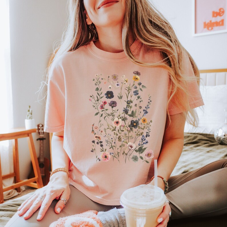 May include: A pink t-shirt with a floral print featuring a variety of flowers in shades of pink, blue, yellow, and white.
