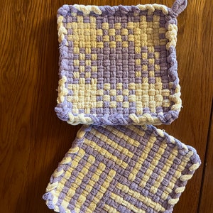 May include: Two square woven pot holders in a purple and cream color pattern. The pot holders have a braided edge.