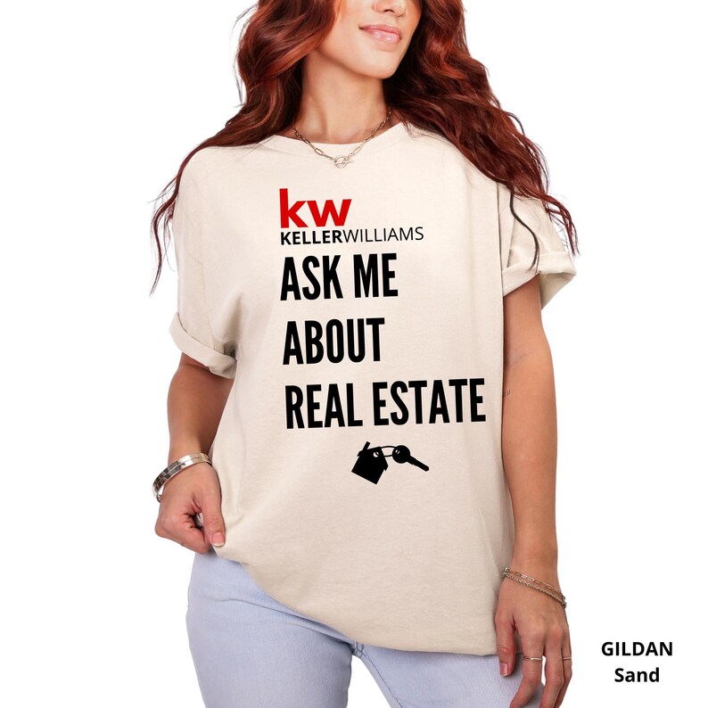 May include: Sand-colored t-shirt with the Keller Williams logo and the text "ASK ME ABOUT REAL ESTATE." A graphic of a house and key is below the text. The shirt is a Gildan brand.