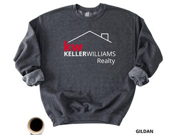 May include: A dark heather grey crewneck sweatshirt featuring the white and red Keller Williams Realty logo and house outline graphic on the chest. The garment has long sleeves with ribbed cuffs and a ribbed neckline, displaying the brand name clearly.