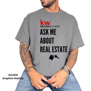 May include: Graphite heather t-shirt with the Keller Williams logo and the text "ASK ME ABOUT REAL ESTATE". The design includes a house and key graphic. The shirt is made of a soft, comfortable material.