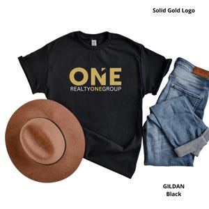 May include: A black short-sleeve t-shirt featuring the gold text ONE REALTYONEGROUP on the chest. The shirt is displayed alongside a brown felt fedora hat and a pair of blue denim jeans with rolled cuffs, showcasing a casual ensemble.